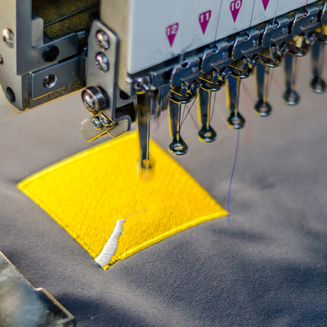 Máquina de bordado industrial computadorizada aplicando linha amarela em tecido escuro. Detalhe da agulha criando um patch bordado de alta definição.